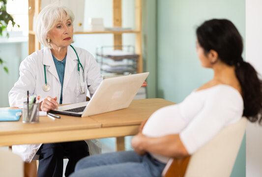 At clinic, pregnant woman is at doctors appointment. Senior medical female specialist and patient sit at table and communicate, interview, anamnesis, patient complaints