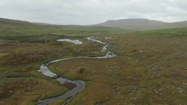 A drone shot pushes in over a lush, remote valley on the Faroe Islands, showcasing the winding river Sand&aacute;, small lakes, and distant hills under a cloudy sky.