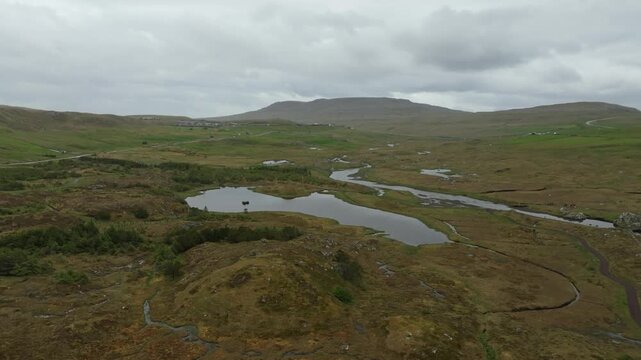 Aerial drone shot panning left over a lush, remote valley on the Faroe Islands, featuring the winding river Sand&aacute;, the tranquil Grunnatj&oslash;rn lake, and a distant village under cloudy skies.