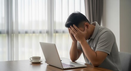 Fototapeta premium A stressed man at his desk holds his head in frustration while working on a laptop. A coffee cup sits nearby. The scene captures work burnout and overwhelming stress in an office setting.