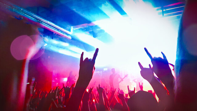Crowd of people with heavy metal devil horn sign mano cornuta hands up at live rock music concert show in spotlight and band group real photo, no ai