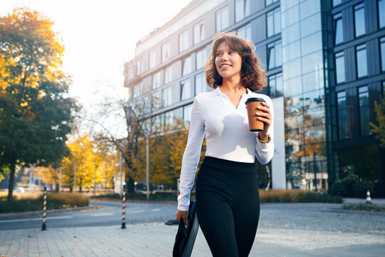 Pretty woman smiling with coffee outside contemporary office building. Attractive woman holding business briefcase in elegant professional style. Urban career concept.