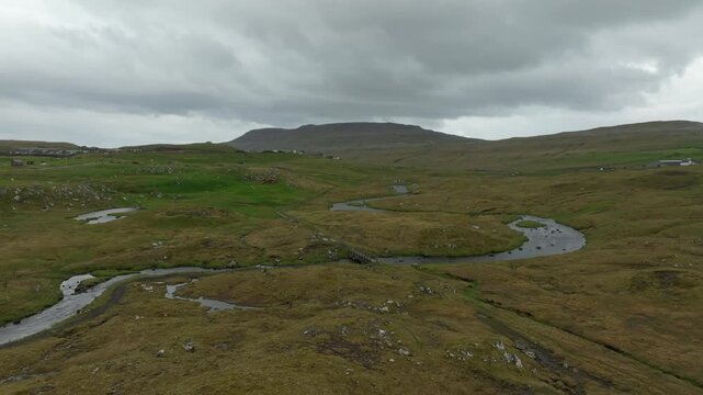 An aerial push in drone shot reveals the winding river Sand&aacute; through a lush, rugged valley with a distant village on the Faroe Islands, under a cloudy sky.