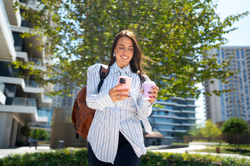 Naklejka premium A woman walks in an urban area while using her phone. She holds a cup in one hand and has a backpack on her back. It is a sunny day with buildings in the background.