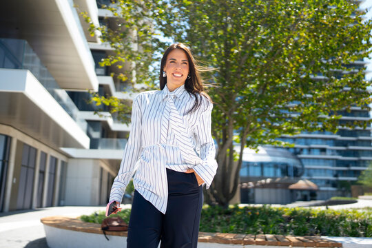 A woman walks on a sunny day near a large building with green trees. She wears a striped shirt and looks happy while holding her phone. The setting shows a clean and bright area.