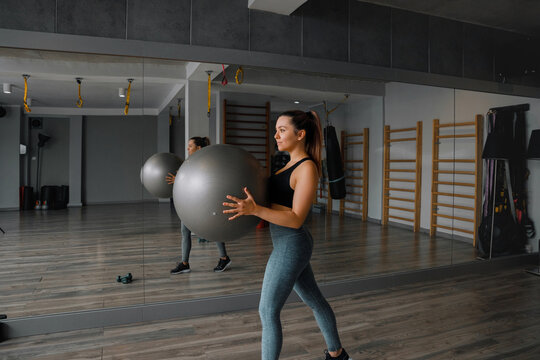Young woman holding fitness ball during energetic gym workout indoors. Pretty woman training core muscles with balanced athletic posture inside studio. Fitness wellness concept.