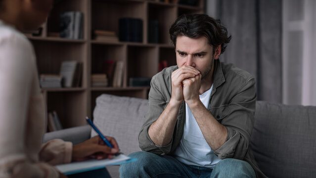 Worried Young Caucasian Man Deep In Thought, Consulting With Therapist Focusing On Mental Health While Sitting In Modern Office Setting. Emotion Reflects Concern And Anxiety Regarding Personal Issues.