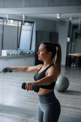 Fototapeta premium Young woman training with dumbbells during energetic indoor fitness exercise routine. Pretty woman lifting hand weights with athletic balanced posture inside studio. Strength wellness concept.