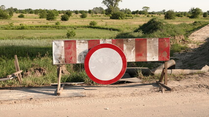 Znak zakazu ustawiony na biało-czerwonej zaporze drogowej. Blokada odcina zniszczoną, polną drogę na tle wiejskiego krajobrazu z zielonymi polami i drzewami w słoneczny dzień © tomo81
