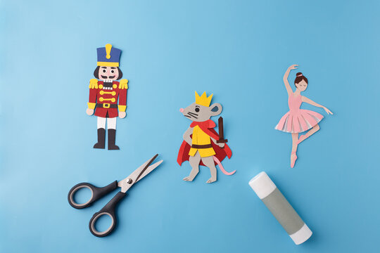 Paper cut nutcracker soldier, king mouse and ballerina with scissors and glue stick on blue background. Flat lay craft composition with copy space. Christmas and Nutcracker ballet concept. 