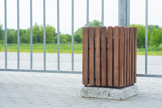 Modern wooden trash bin with vertical slats on concrete base in urban public space. Street furniture, city infrastructure and waste management equipment on paved sidewalk near metal fence.