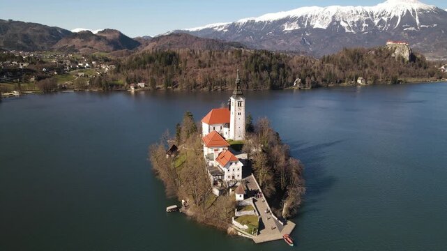 Aerial view of the iconic pilgrimage church on the island in Lake Bled, surrounded by deep blue water and snow-capped mountains in Bled, Radovljica, Slovenia.