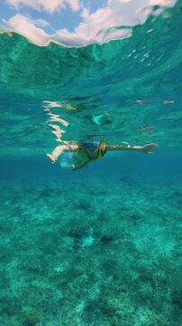 Vertical Screen: Woman in yellow life vest and blue snorkel mask glides through crystal clear turquoise ocean waters above coral reef floor with cloudy sky visible through rippling surface above.