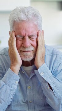 Elderly caucasian man with a beard massaging his temples due to a severe headache, feeling stressed and tired while experiencing intense pain and discomfort in his head at home