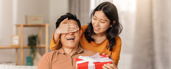 Obraz premium Joyful Emotion. Happy Young Asian Woman Surprising Man With Gift Box While Covering His Eyes, Celebrating A Special Occasion Together At Cozy Home Setting. Family Concept.