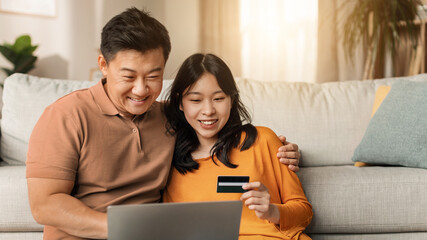 Fototapeta premium Smiling Asian Father And Daughter Engaged In Online Payment Activity Together At Home, Sitting On Sofa With Laptop And Credit Card, Sharing Joyful Moment In Cozy Living Room. Family Concept.