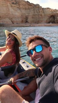 Young smiling couple having fun filming a selfie video while kayaking near a stunning cliff coastline. Joyful boyfriend and girlfriend enjoying their summer vacation with an outdoor water sport