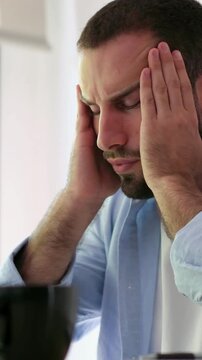 Exhausted young man feeling stressed while working on a computer at his office, massaging his temples and head to relieve a painful migraine and pressure from overworking and fatigue