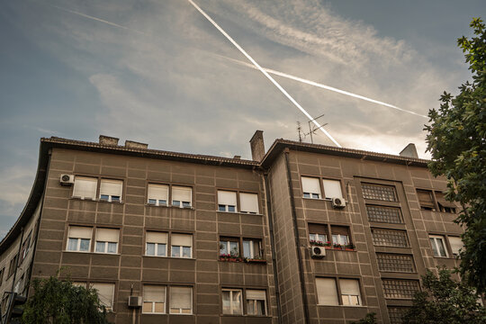 Residential apartment block in Belgrade, Serbia, showing a concrete facade, window shutters, air conditioners, balconies, and evening sky with aircraft contrails above the urban housing complex.