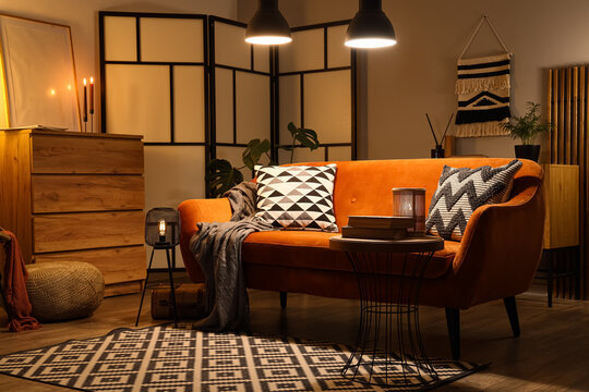 Interior of living room with folding screen, commode, glowing lamps and soft plaid on orange sofa in evening