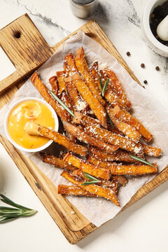 Wooden board with tasty cooked sweet potato, grated Parmesan cheese, rosemary and bowl of sauce on marble background