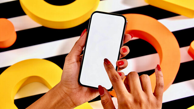 Top view of woman hands holding smartphone with blank white screen on geometric black and white striped background with yellow and orange 3D shapes.