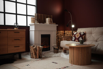 Obraz premium Interior of living room with sofa, commode, coffee table, fireplace and firewood in basket near burgundy wall