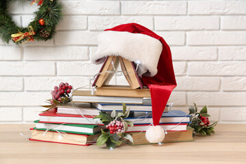 Fototapeta premium Christmas tree made of books, berries, glowing lights and Santa hat on table near white brick wall