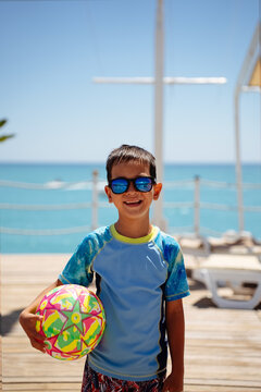 Happy young boy in sunglasses holding colorful beach ball on wooden seaside pier with turquoise ocean background. Vertical shot of summer vacation mood, family travel and resort leisure concept.