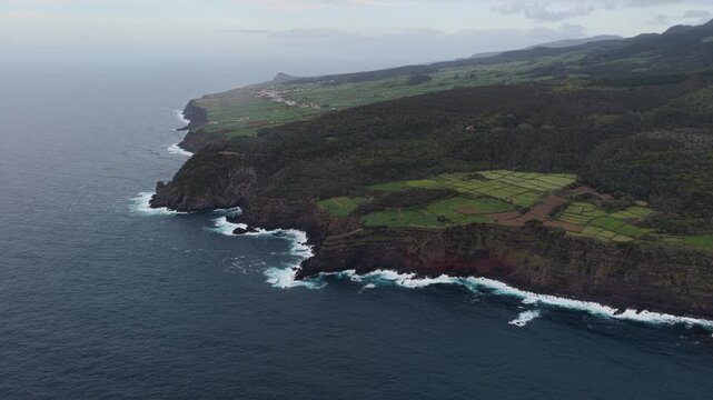 Aerial view of the rugged coastline near Miradouro da Ponta do Queimado, featuring dramatic volcanic cliffs, green fields, and the Atlantic Ocean in Serreta, Acores, Portugal.