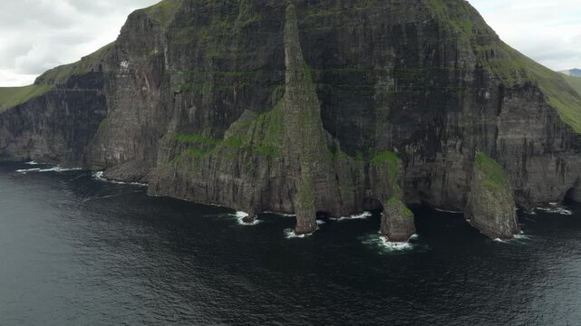 Aerial drone shot showcasing the rugged coastline and majestic cliffs called Tr&oslash;llkonufingur (The Witch&rsquo;s Finger) on the Faroe Islands, with the camera performing a pull back, boom up, and pan left.