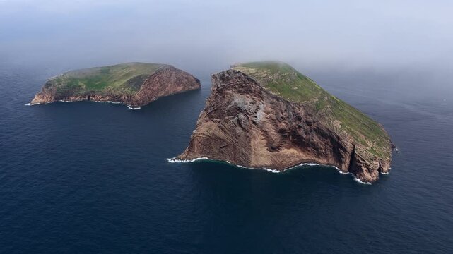 Aerial view of the Ilheus das Cabras islets featuring steep rocky cliffs and grassy slopes surrounded by the deep blue Atlantic Ocean in Azores, Portugal.