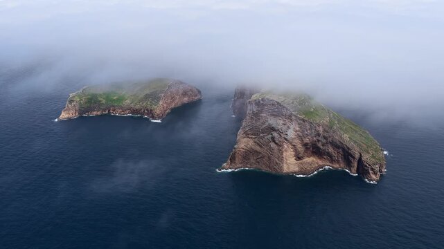 Aerial view of Ilheus das Cabras, two rocky islets rising from the Atlantic Ocean, partially shrouded in low-hanging clouds, Azores, Portugal.