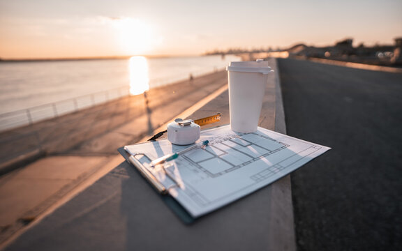 Architectural blueprint, measuring tape,takeaway coffee rest on waterfront promenade during golden hour. Coastal skyline and sunset create inspiring atmosphere for urban planning and design decisions.