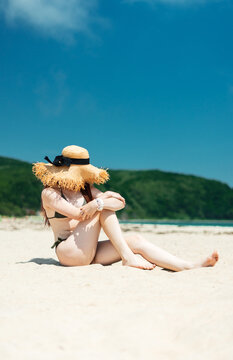 Woman in bikini and straw hat sitting on soft sandy beach under blue sky, enjoying sunlight, peaceful ocean atmosphere. Summer vacation, tropical escape, relaxation, freedom, serene coastal lifestyle.