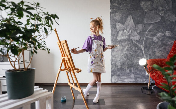 Little girl painting on canvas at home studio, holding palette and brush. Creative hobby, art education and child development concept. Cozy interior with natural light and plants.