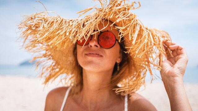 Young stylish woman wearing straw hat on beach. Summer fashion, trendy accessories and travel lifestyle concept. Bright sunlight and tropical background create relaxed vacation mood