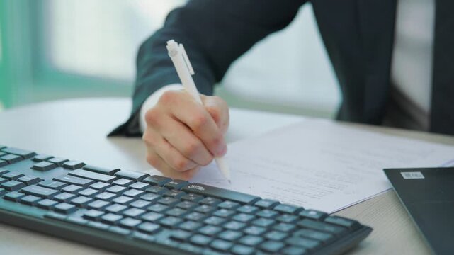 A close-up of a person's hand writing with a pen on a document beside a laptop keyboard in a bright modern office environment.