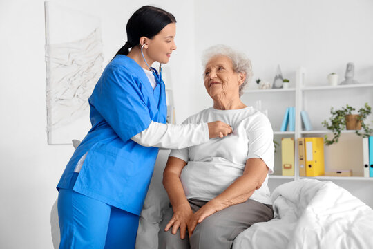 Female nurse hearing senior woman with stethoscope in bedroom