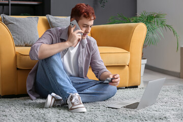 Fototapeta premium Handsome young man with credit card, mobile phone and modern laptop sitting on floor near sofa while shopping online
