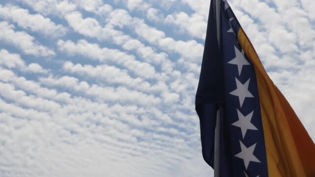 The flag of Bosnia and Hercegovina waving in the wind on a flag pole with the blue sky and white clouds behind it. 
