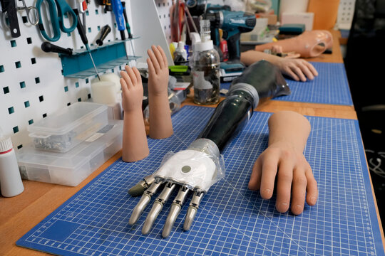 Tools hang above the work surface used to create and customize prosthetics. Various artificial hands and a partial arm rest on a gridded workbench in a well lit workshop