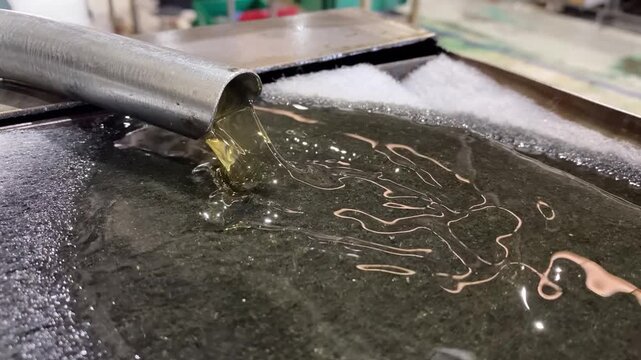  Close-up video of industrial lubricating oil flowing from a metal pipe into a storage tank inside a manufacturing facility. The footage highlights fluid transfer, lubrication