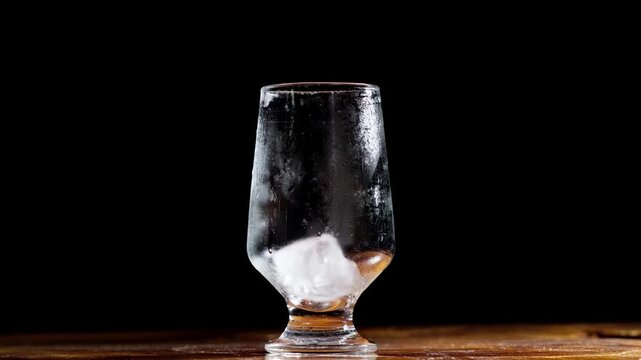 Metal tongs gently place ice cubes into chilled glass centered in black studio background