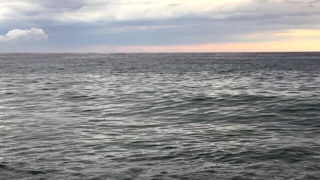 Rain falling over the sea beneath dramatic storm clouds. Gentle ocean waves ripple under a dark overcast sky as distant rainfall creates a moody coastal seascape before the storm.