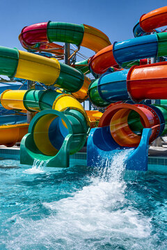 A colorful water slide complex with multiple tubes and splash pools on a sunny day. The water is clear blue, and the slides are various bright colors like yellow, green, orange, and blue.