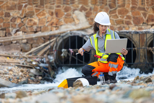 Environmental engineer using laptop to analyze water samples near industrial drainage, Professional female scientist testing water quality at polluted site with field equipment