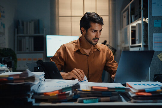 Overworked young Latin businessman working late on document in large stack of paperwork files at office night. Tax audit deadline, accounting stress, corporate burnout, urgent business concept.