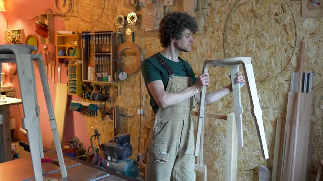 Private production of wood products, cheerful focused man carpenter examines semi-finished chair, craftsman holding stool and precisely checking quality of wooden elements, crafter proud of new item.