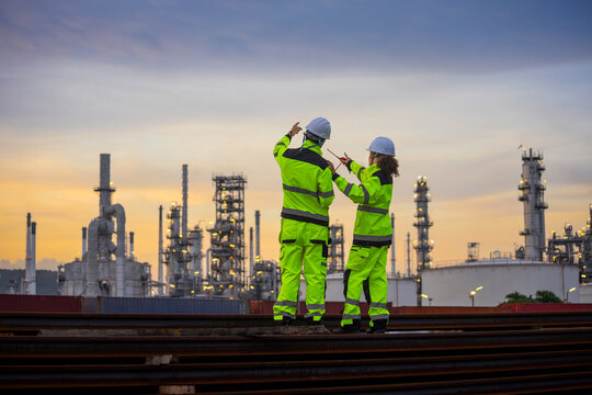 Industrial engineers discussing project progress at oil refinery plant during sunset, Professional construction team in safety gear inspecting petrochemical facility with radio communication